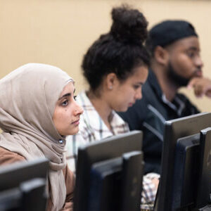 Three Students sit in a row. There are computers on the table in front of them