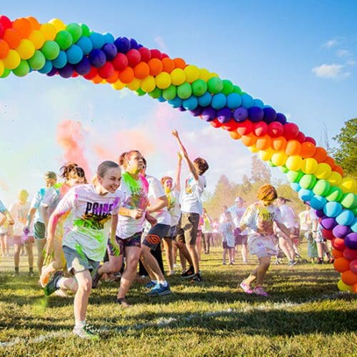 students throw color powder in the air while running under a rainbow balloon arch