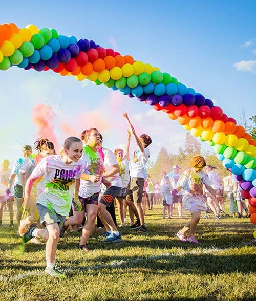 students throw color powder in the air while running under a rainbow balloon arch