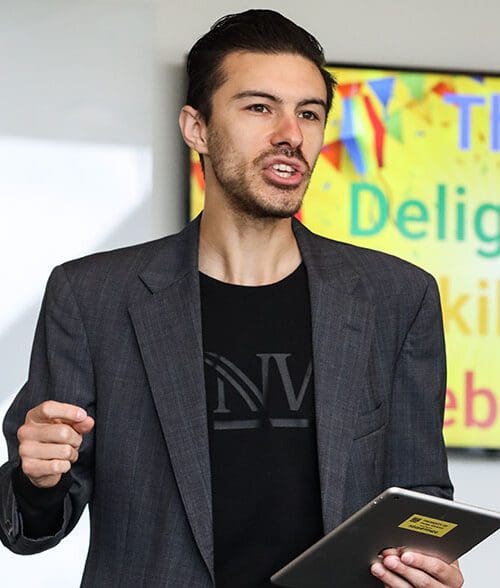 Nathan Elizalde stands with a tablet in his hand. There is a screen behind him that reads "The Delightful Skill of Celebration"