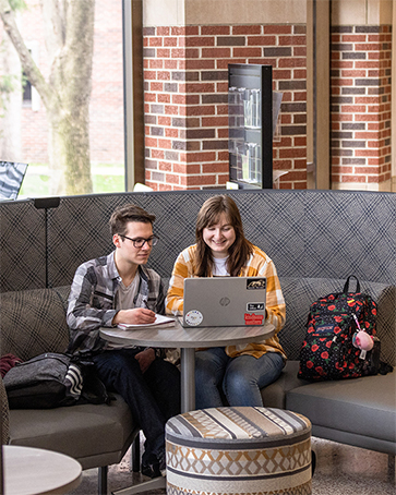 Two students sit in a booth and look at a laptop on the table in front of them.