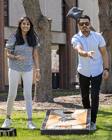 Two students playing cornhole, the bag is mid air, coming toward the camera.