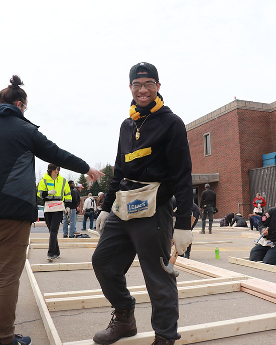 An honors college student smiles while holding a hammer. He is standing on top of a framed wall.