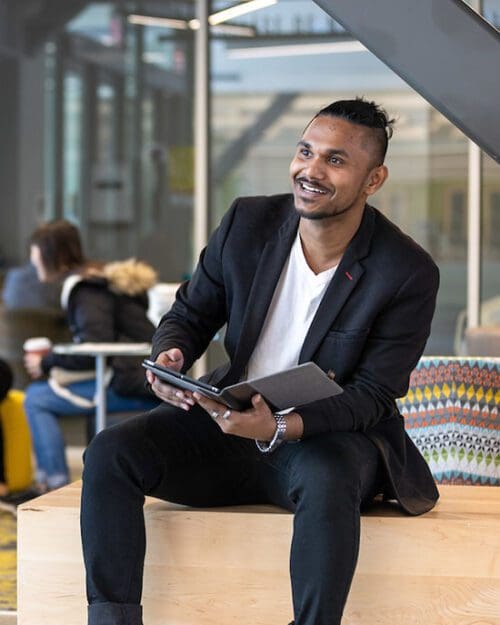 A student sits on a couch. He is looking off to the left of the camera. There is a folder in his hands.