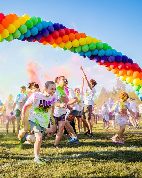 Students run under a rainbow balloon arch. There is powdered color going in the air behind the arch.