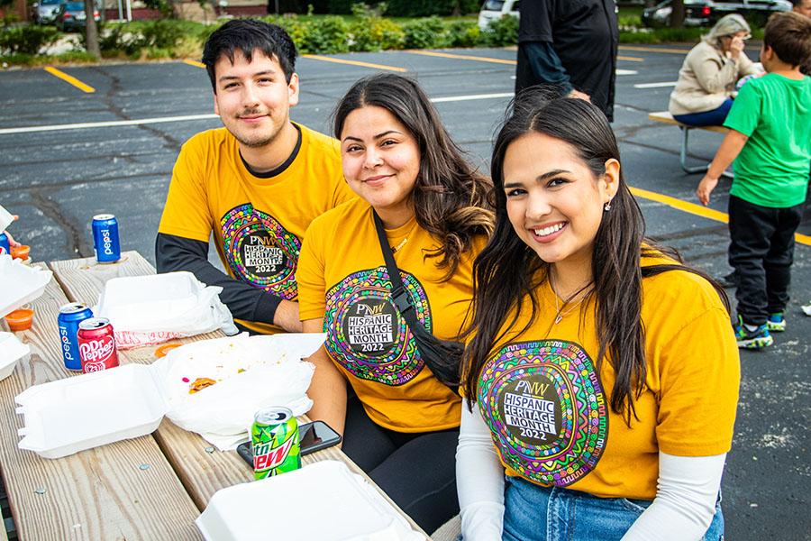 Three students in matching yellow "Hispanic Heritage Month" t-shirts pose together while sitting at a picnic table. There are to-go containers on the table in front of them.