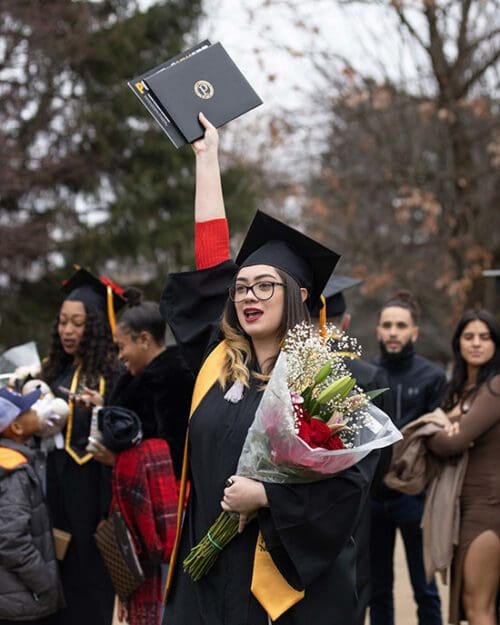 A PNW graduate with flowers holds her diploma high.