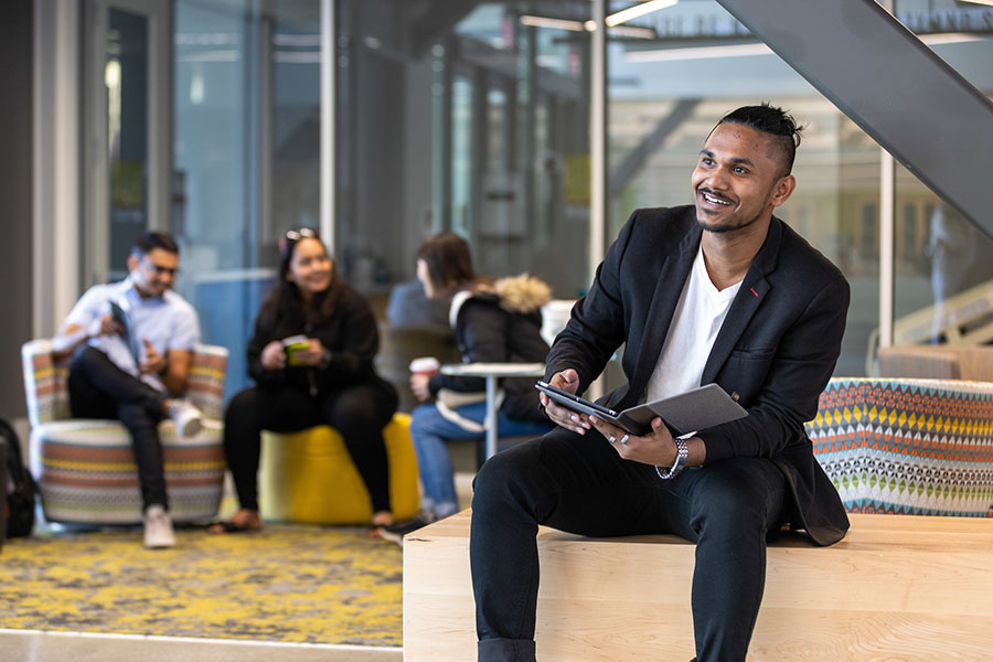 A student sits in the Nils K. Nelson Bioscience and Innovation Building. He is holding a folder and there are three more students sitting on a couch in the left background.