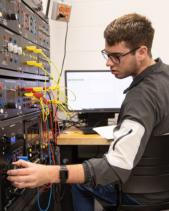 Student sits in a chair and leans to the left to work on electrical wiring. There is a table with a computer in front of him.