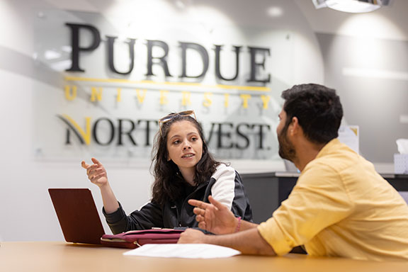 Two students sit at a table. The student to the left has a laptop on the table in front of them. The PNW logo is on the wall behind them.