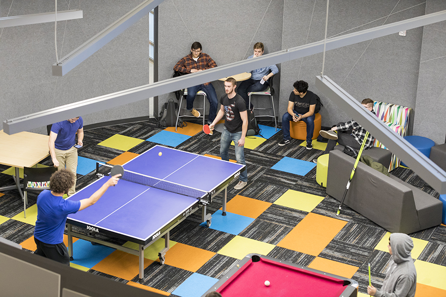 Students sit around a ping pong table. Two students are actively playing ping pong.