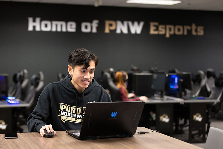 A student sits at a table and looks at an open laptop in front of them. The wall behind them says "Home of PNW Esports" and there are rows of computers and several students sitting at those computers in the background.