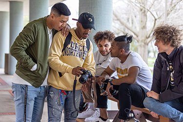Three students sit on a picnic table, two students stand next to them. One student is showing the group a camera.