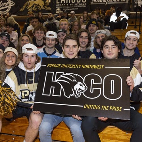 Students sitting in bleachers. The students in the front row are holding a black homecoming sign.