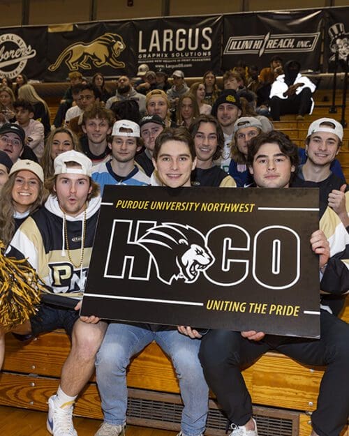 Students sitting in bleachers. The students in the front row are holding a black homecoming sign.