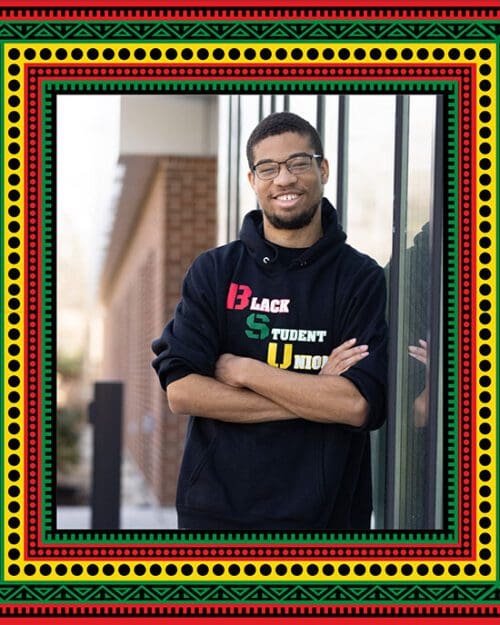 PNW student David Bolton stands on campus wearing a Black Student Union t-shirt. A white PNW logo on a black background. There is a red, yellow, and green traditional African-style border around the image.