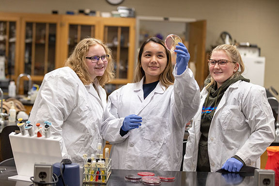 Three students stand together wearing white lab coats and blue, plastic gloves. The student in the middle is holding a petri dish in the air.