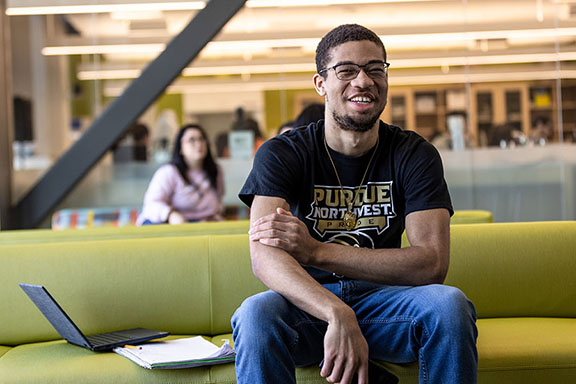 A student sits on a couch and smiles. There is a laptop and notebook on the couch next to them and many students sitting and walking around in the background of the photo