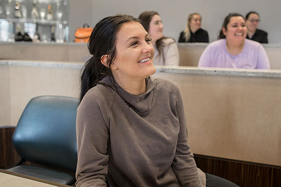 A student sits in a classroom and smiles while looking off to the right. There are several students sitting in the rows behind the student in focus.