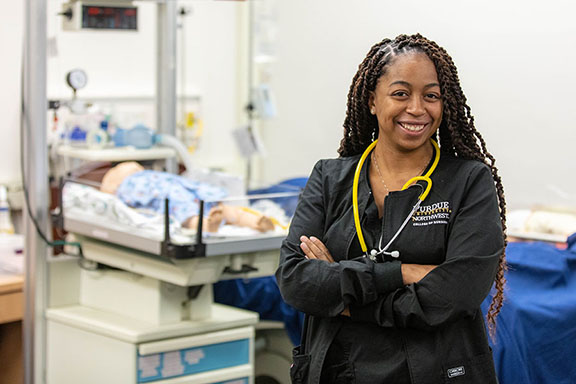 A student stands with their arms folded in a nursing lab room. They are wearing scrubs and have a stethoscope draped around their neck.