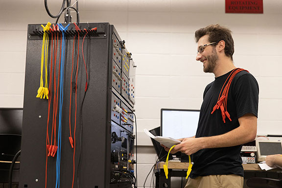 A student stands in front of a tower of electrical boxes, they have red cords draped over their shoulder and are smiling.