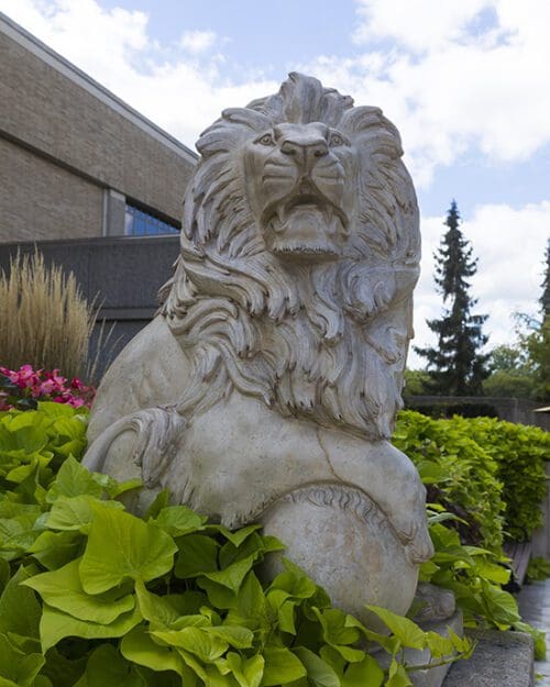 A lion statue with greenery around the bottom and a building in the background