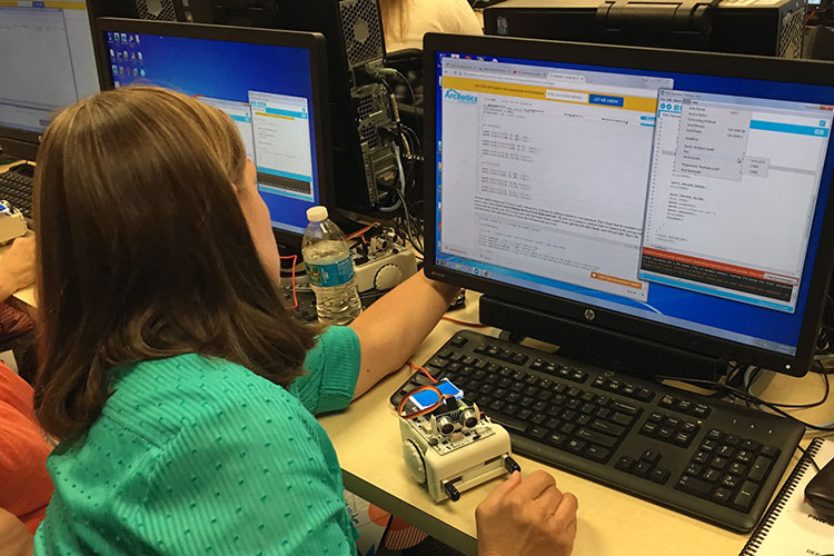 A GenCyber Camp participant codes at a computer with a robot in front of her.