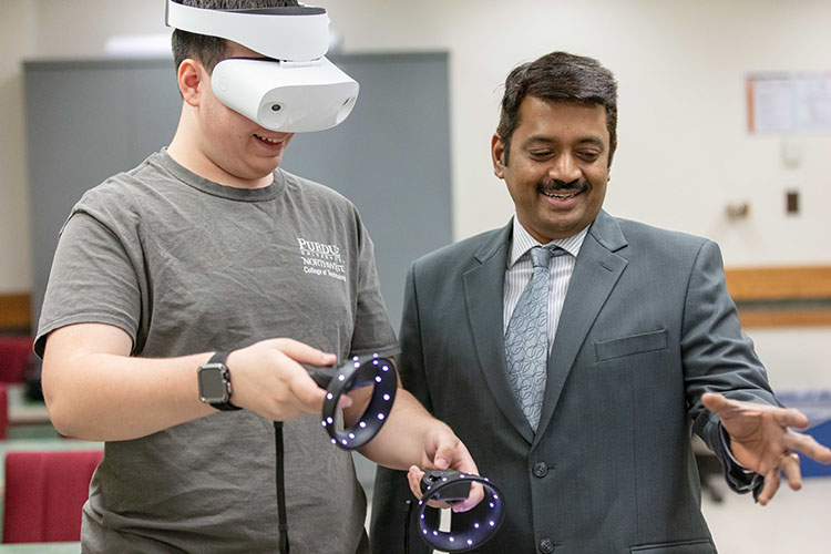 Magesh Chandramouli smiles and points while standing next to a student who is wearing a VR headset