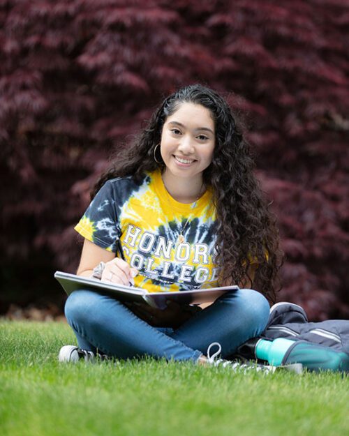 A student sits cross legged in the grass. They have a notebook open on their lap.