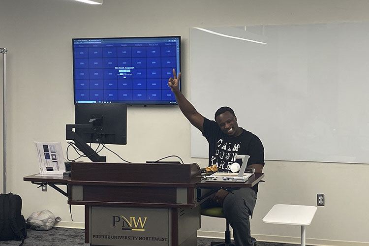 A student smiles and holds a "peace" sign in the air while sitting in front of a computer