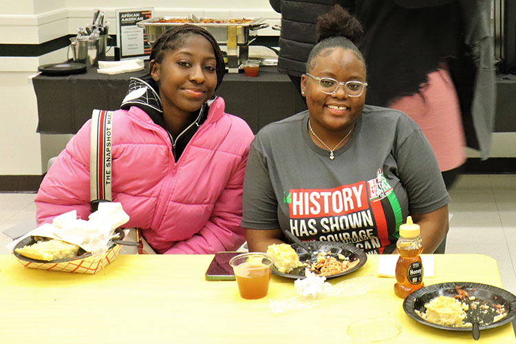 Two students sit at a table. There are plates of food sitting on the table in front of them.