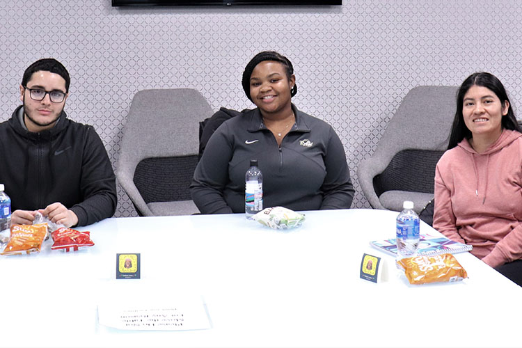 Three students sit together at a white table. There are snacks on the table in front of them.