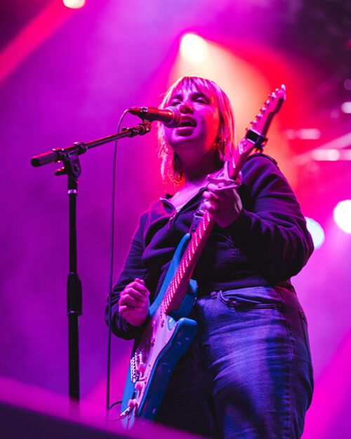 Lili Trifilio, the lead singer of Beach Bunny, stands in front of a microphone and sings while playing the guitar