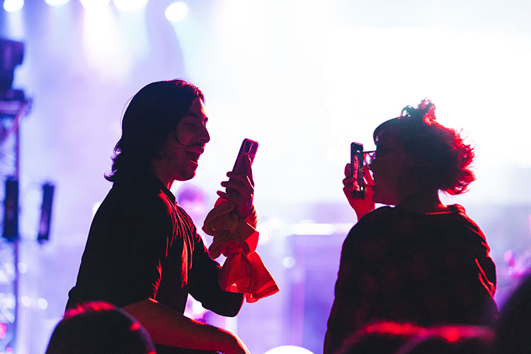 Two audience members sit on the shoulders of their friends. They are holding their phones up to record.