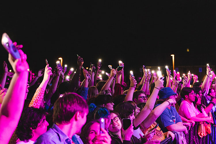 Audience members are shown in pink light holding up their cellphone flash lights