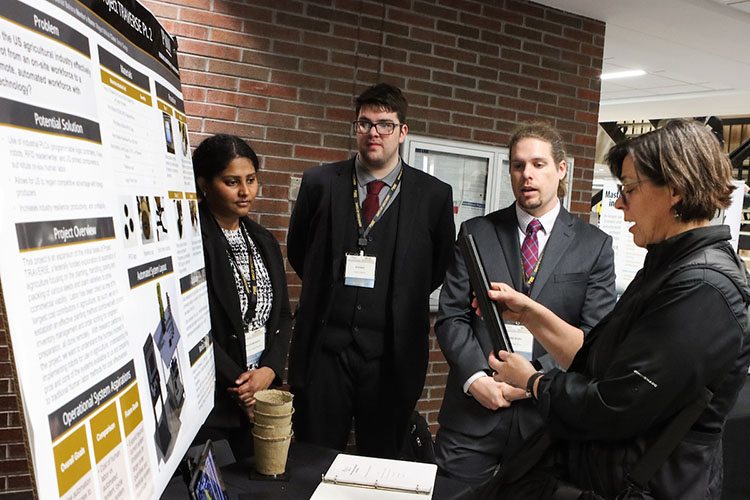 Three students stand by their poster presentation. Another person stands in front of the poster, looking at it.
