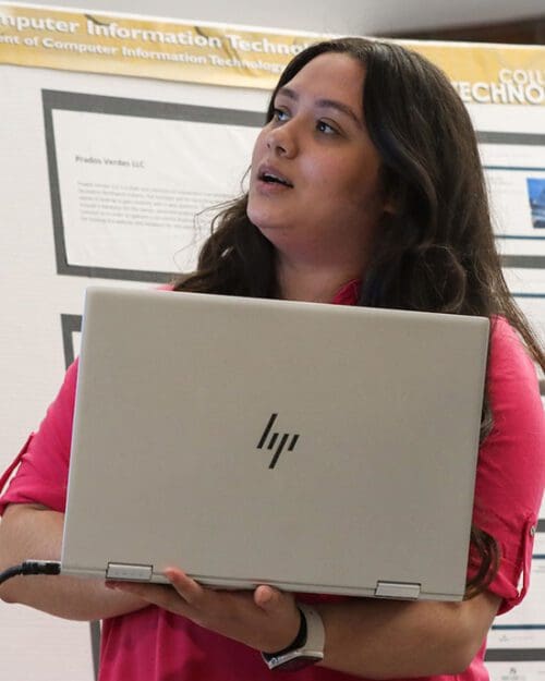 A student stands in front of a printed poster presentation. They are holding a laptop and looking off to the side.