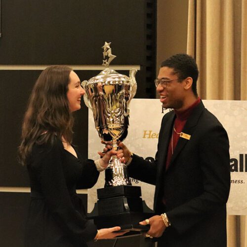 Two students stand facing each other. They are holding a trophy.