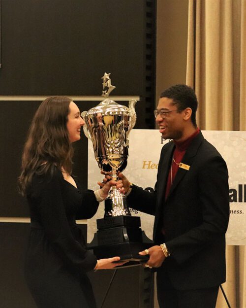 Two students stand facing each other. They are holding a trophy.