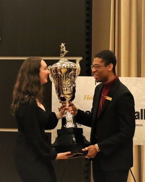 Two students stand facing each other. They are holding a trophy.