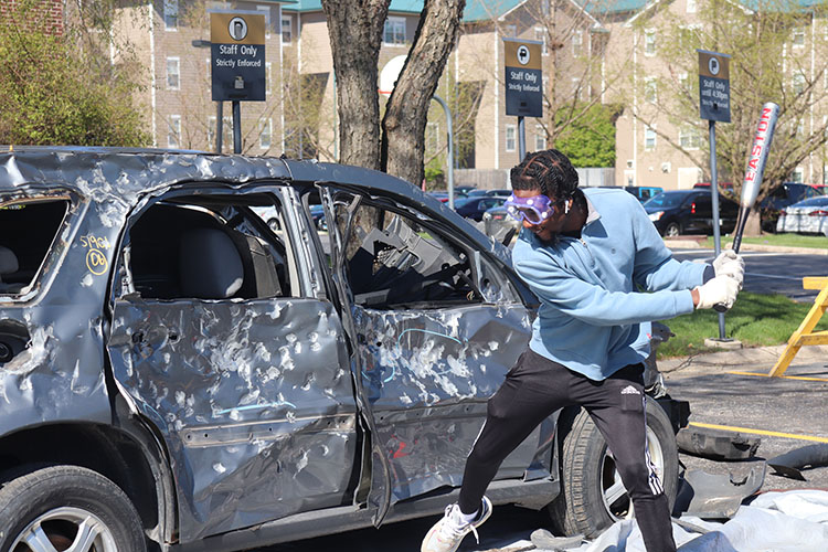A student holds a baseball bat. There is a dented car in front of them