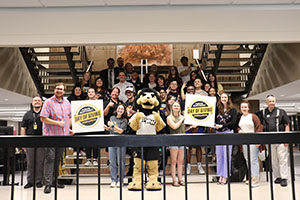 A group of people stand on a stair landing and hold two day of giving signs. Leo the Lion is standing in the middle of the group.