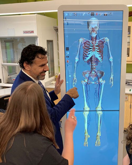 Two people look at an anatomage table that is featuring a skeleton