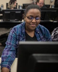 A student sits behind a computer. They are looking at the monitor and smiling