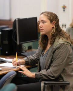 A student sits at a table with a row of computers on it. They are writing on a piece of paper and looking off to the left