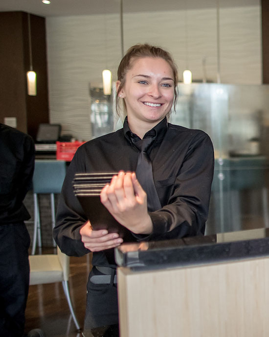 A student in a black dress shirt and tie stands behind a podium. They are smiling and holding a stack of menus