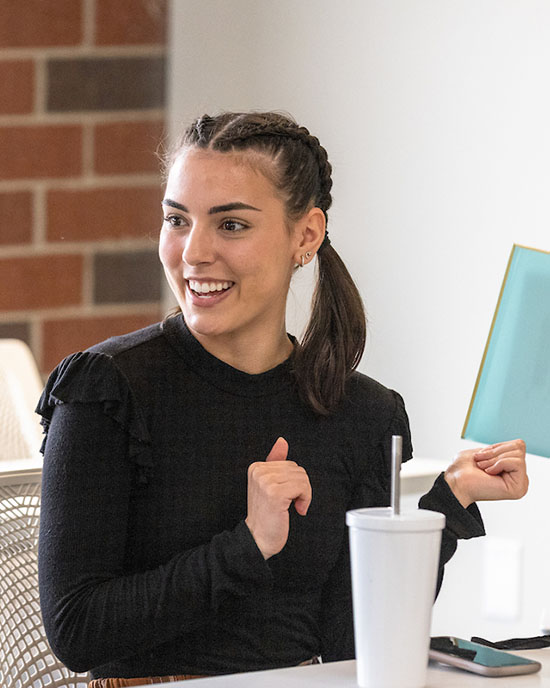 A student in a black shirt sits at a table. They are looking to the left and smiling. They are holding a blue folder with their right hand