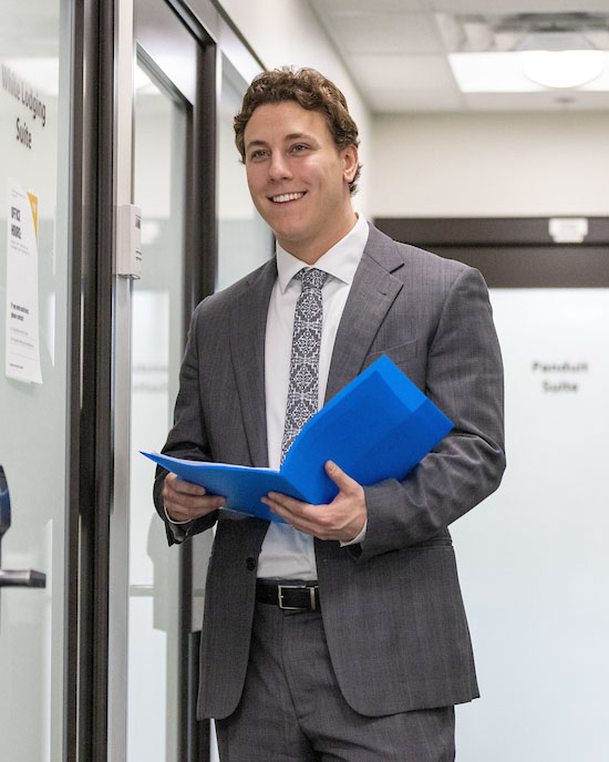 A student stands in a gray suit. They are holding a blue folder