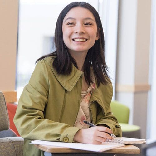 A student in a green jacket and floral shirt sits at a desk. Their hand is resting on an open notebook on the desk. They are holding a pen.