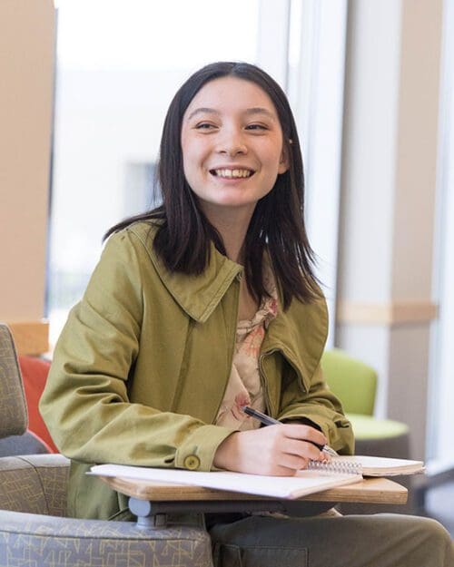 A student in a green jacket and floral shirt sits at a desk. Their hand is resting on an open notebook on the desk. They are holding a pen.